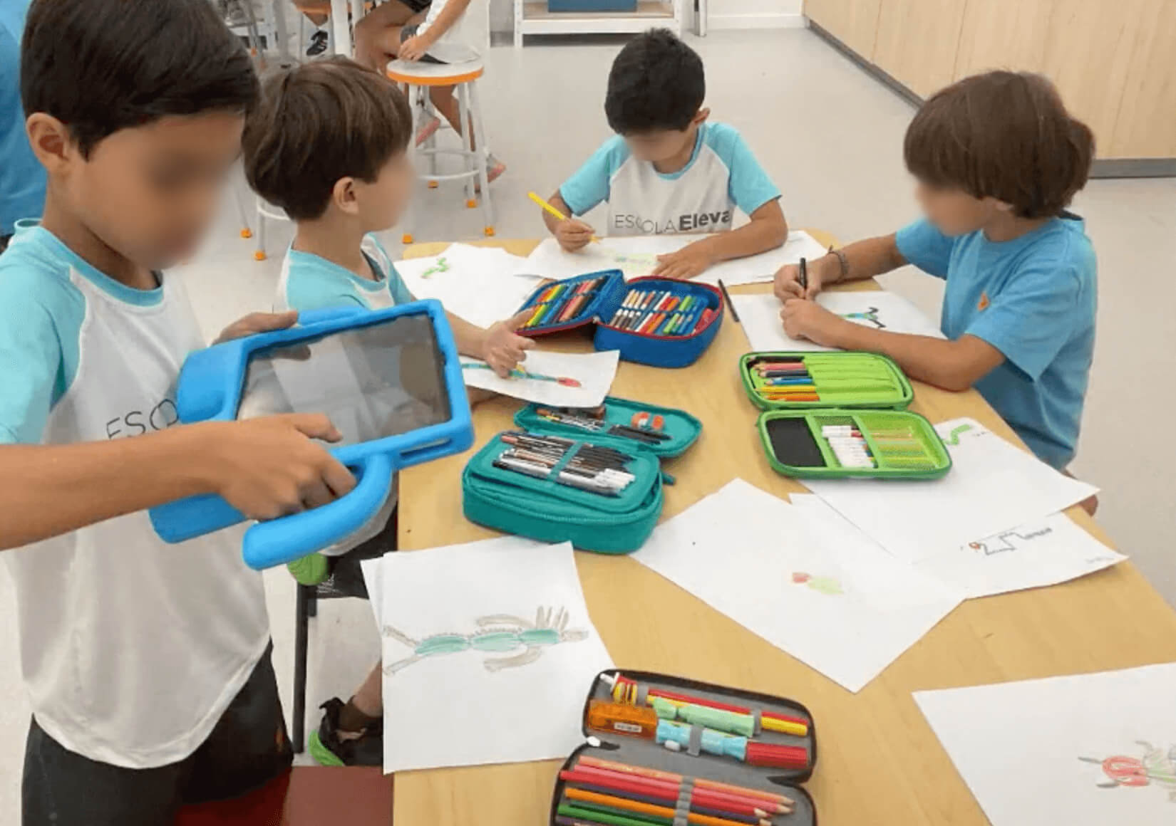 A group of second-grade students at Escola Eleva are engaged in various activities around a table. One boy in the foreground is holding a tablet with a blue protective case. Other students are drawing with colored pencils and markers, their pencil cases open and filled with art supplies. The students are wearing light blue and white Escola Eleva t-shirts. The classroom setting is bright and spacious, with stools and tables in the background.