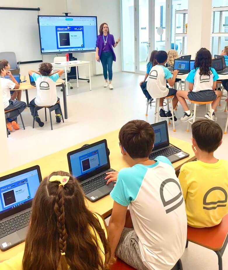 A classroom scene featuring third-grade students at Escola Eleva. The students are seated at tables, each with a laptop, while a teacher stands at the front of the room. The teacher is wearing a purple jacket and is speaking to the class, with a large digital screen behind her displaying a Book Creator page titled 'About me.' The students are wearing school uniforms in blue and white or yellow and white, with some students focusing on their laptops and others listening to the teacher. The classroom is bright and modern, with large windows allowing natural light to enter.