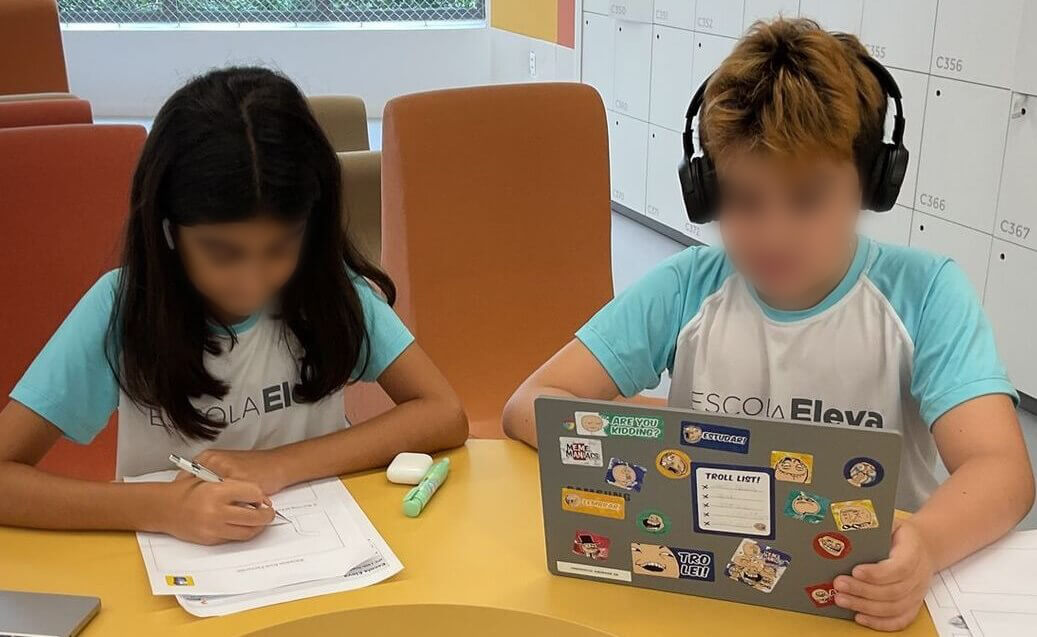 Two fifth-grade students at Escola Eleva are working together at a table. The girl on the left, with long dark hair, is focused on writing on a worksheet. The boy on the right, wearing headphones and with light brown hair, is working on a laptop covered in colorful stickers. Both students are wearing Escola Eleva uniforms with light blue sleeves. The background features lockers and large windows that allow natural light into the room.