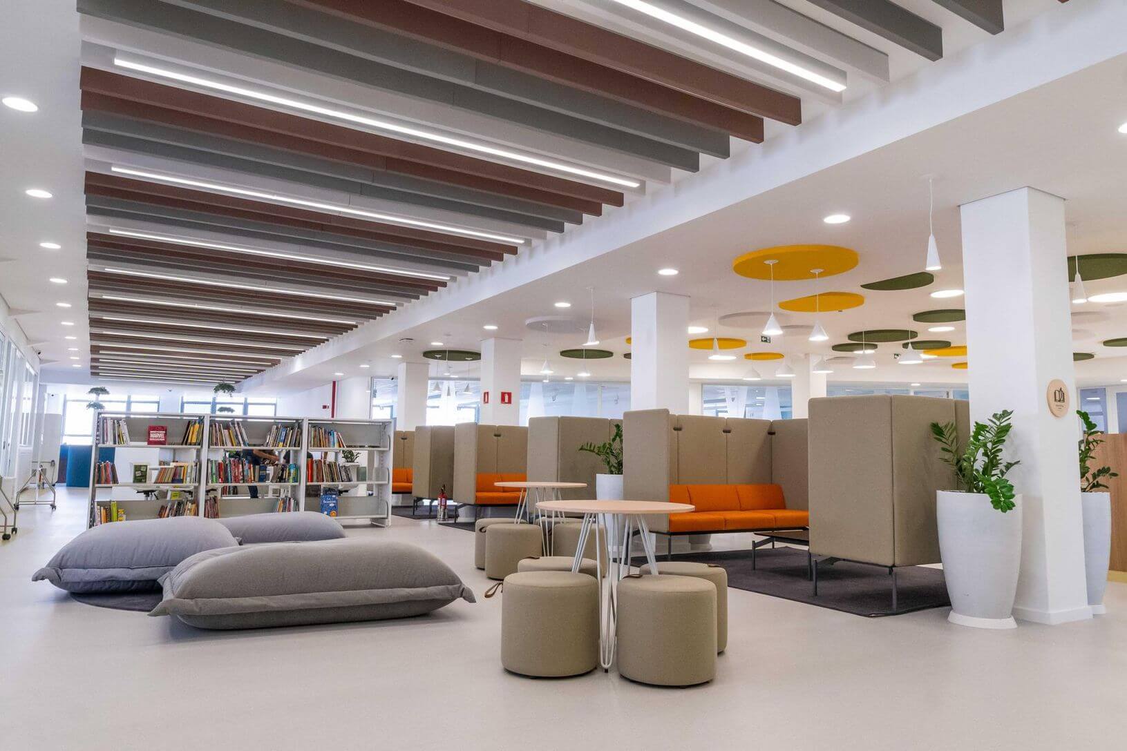 Inside Escola Eleva - a modern, open-concept school library or common area with a clean and minimalist design. The ceiling features alternating colored panels in shades of brown, gray, and white, along with circular light fixtures in green and yellow. The space includes various seating options, such as large gray cushions on the floor, beige stools, and enclosed seating pods with orange cushions. There are several white bookshelves filled with books, and large potted plants add a touch of greenery. The area is brightly lit with natural light coming in through large windows.