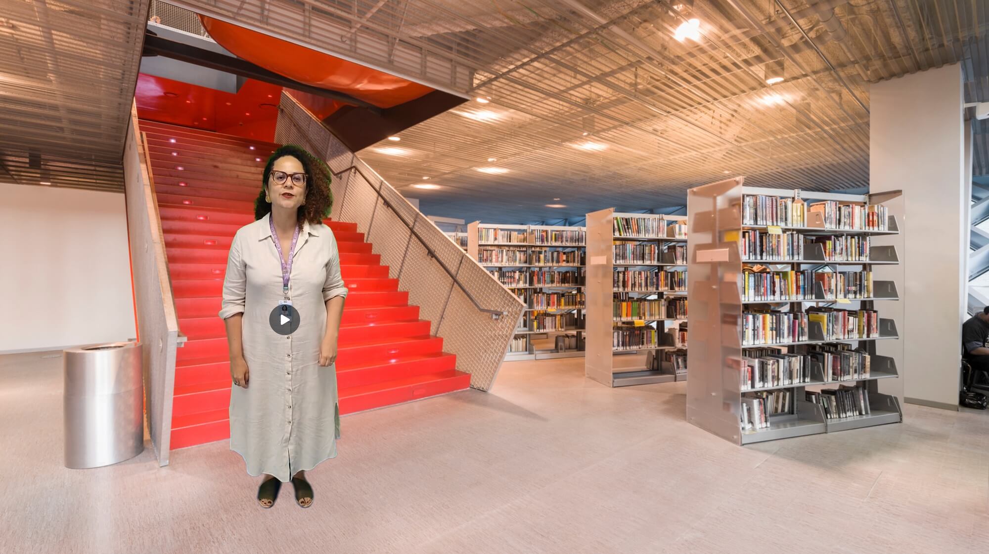 A woman stands in a modern library, next to a striking red staircase. She is wearing a light-colored dress and has a lanyard around her neck. Behind her, the library is filled with shelves of books. The ceiling features an industrial design with exposed elements. The woman appears to be giving a presentation or a tour, indicated by her confident stance and the play button overlay on her image.