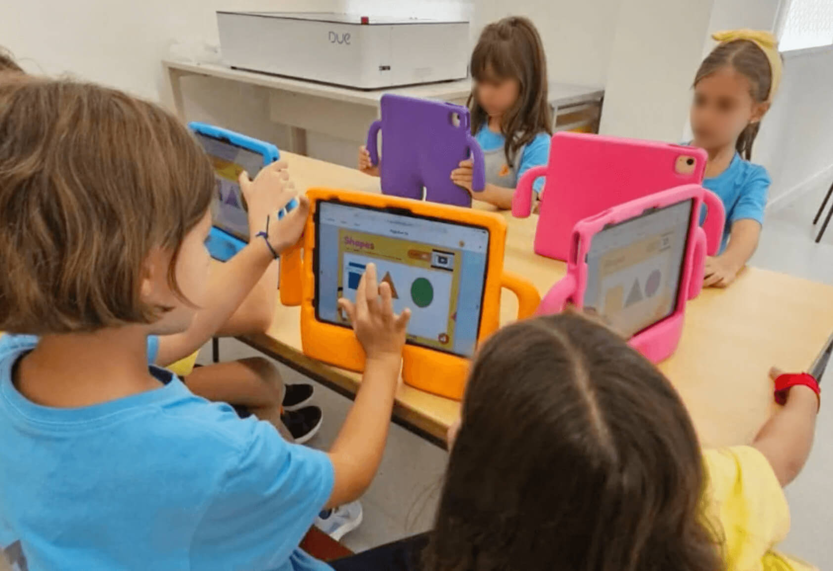 Several young students are seated around a table, each using a tablet with colorful protective cases in blue, orange, pink, and purple. The children are engaged with Book Creator on their screens, working with shapes and other learning materials. The students are wearing light blue shirts, and one girl has a yellow headband. The classroom setting is bright and modern, with a printer and other materials in the background.